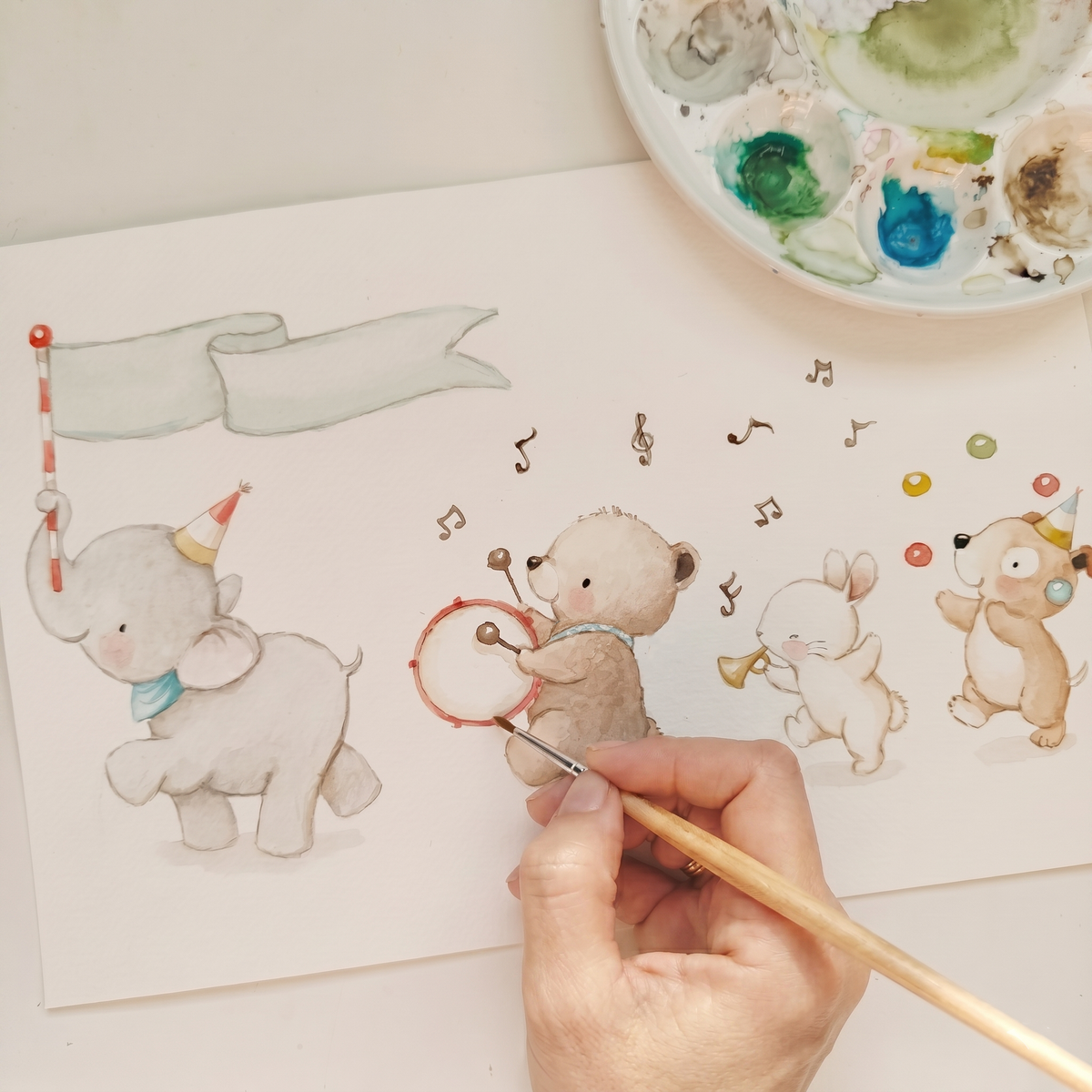 Artist hand painting the original Animal Music Band watercolor illustration of elephant, bear, bunny, and puppy on paper next to a watercolor palette